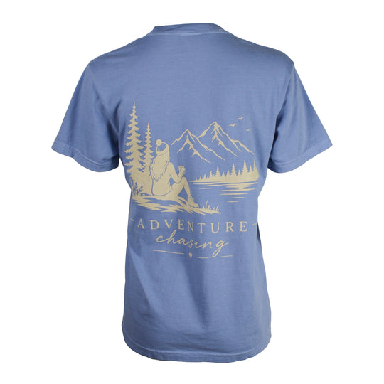 A blue short-sleeve t-shirt shown from the back, featuring a light beige graphic of a person sitting by a lake with mountains and trees in the background, and the words 'ADVENTURE chasing' printed below.